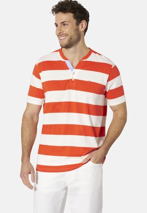Lächelnder Mann mit lockigem Haar trägt ein kurzärmeliges rot-weiß horizontal gestreiftes Shirt und weiße Hose, Hand in der Tasche, schaut nach links.