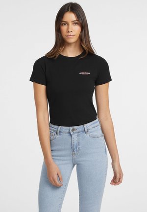 Black cotton t-shirt with short sleeves, crew neckline, and a small red logo on the chest. Paired with light blue high-waisted jeans.