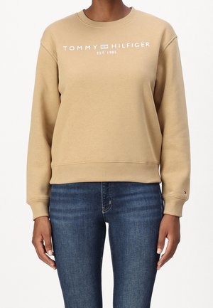 Mujer que lleva una sudadera color beige de Tommy Hilfiger con el logo y el texto "EST. 1985", combinada con jeans ajustados de color azul oscuro sobre un fondo blanco.