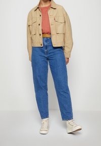 Beige zkrácená bunda s dvěma předními kapsami přes rustikální tričko, doplněná o džínové kalhoty s vysokým pasem a bílé tenisky.