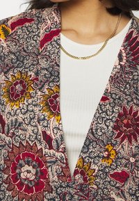 Kobieta ubrana w złoty naszyjnik, biały prążkowany top oraz kolorową kurtkę w kwiatowy wzór z czerwonymi, żółtymi i granatowymi detalami.