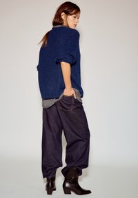 Oversized blauwe gebreide trui met opgerolde mouwen, laagjes boven een grijs shirt, gecombineerd met losse donkere denim jeans en zwarte enkellaarzen.