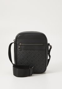 Black crossbody bag with textured logo pattern, leather accents, and adjustable fabric strap. Features a zippered front pocket.