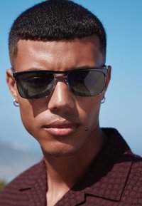 Occhiali da sole rettangolari neri con lenti scure e dettagli argentati. Primo piano con pelle liscia e capelli corti e strutturati.