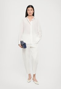 Hvit, semi-transparent skjorte med lange ermer, kombinert med løstsittende hvite bukser. Holder en strukturert blå clutch. Beige spisse hæler.