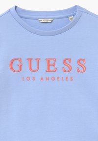 Jasnoniebieski sweter z okrągłym dekoltem. Posiada wyraźny, haftowany napis "GUESS" w kolorze koralowym oraz mniejszy napis "LOS ANGELES" poniżej. Miękka tekstura materiału.