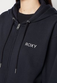 Sweat à capuche zippé bleu marine foncé en tissu doux, doté d'une capuche à double couche, d'un logo "ROXY" brodé et d'une texture lisse.