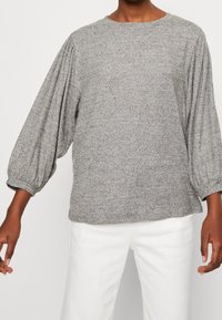 Persona vestita con una blusa grigia a maniche lunghe con maniche a palloncino arricciate e pantaloni bianchi, mostrata dalle spalle a metà coscia.