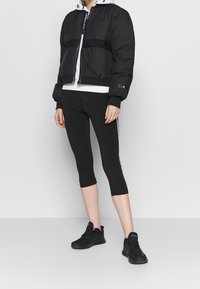 Giacca a vento nera con cappuccio, polsi elastici e strisce nere; abbinata a leggings capri neri e scarpe da ginnastica nere.
