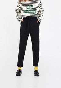 Chemise rayée à manches longues avec texte vert, pantalons noirs taille haute avec poches, chaussettes jaunes et mocassins noirs. Style décontracté, coupe relax.