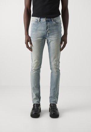 Mann trägt hellblaue Slim-Fit-Jeans, schwarze Loafers und ein schwarzes Hemd, steht vor einem schlichten hellen Hintergrund.