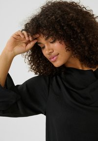 Sort bluse med draperet udskæring og brede ærmer, lavet af et glat stof. Teksturen virker blød og let skinnende.