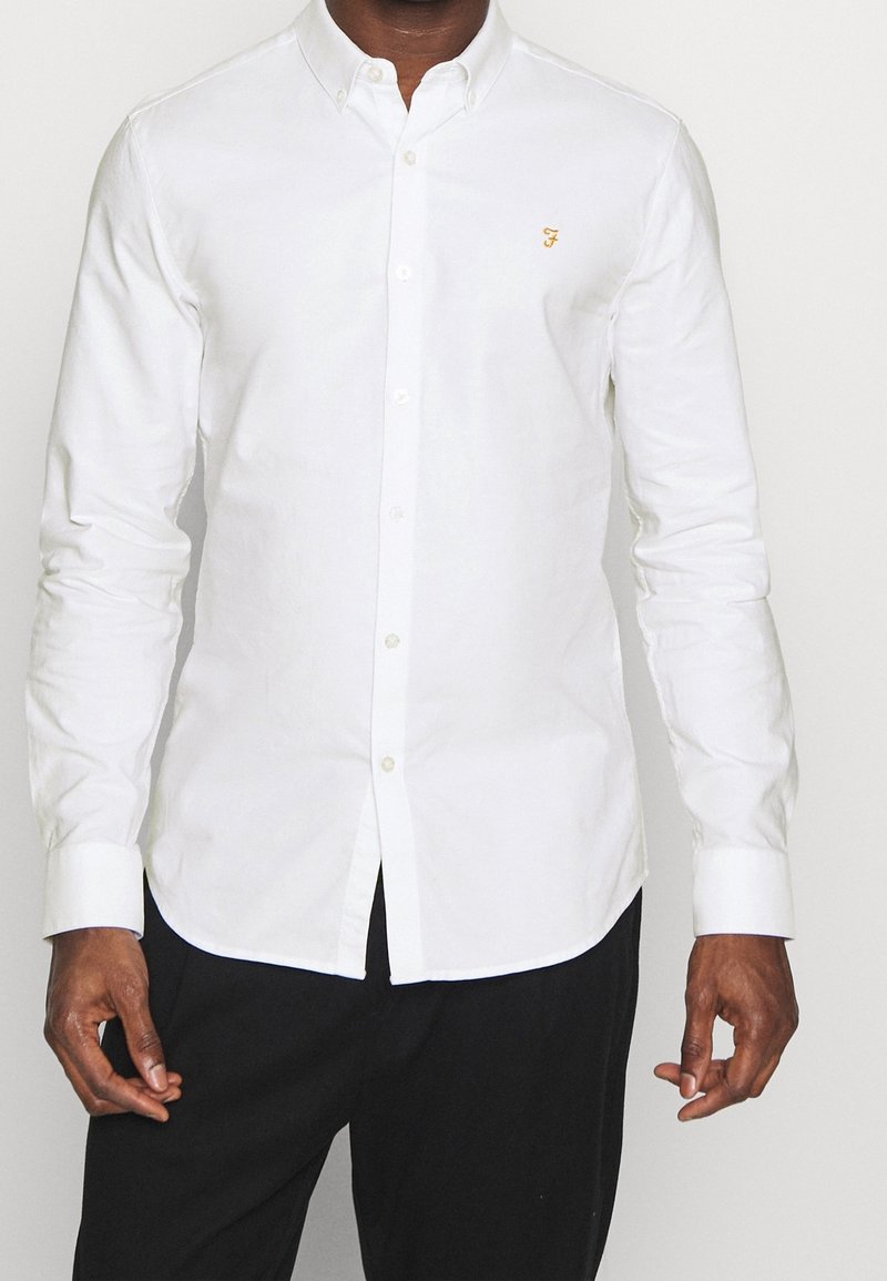 Muž stojící proti jednobarevnému pozadí, oblečený v bílé, na tělo přiléhající košili s dlouhými rukávy a černých kalhotách.