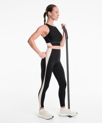 Un crop top noir et des leggings taille haute avec une bande latérale crème. Le modèle tient une barre d'exercice noire et porte des baskets claires.