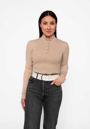 Kvinde med glat, mørkt hår iført en beige ribbet sweater med knapper og sorte jeans med et hvidt bælte, stående mod en hvid baggrund.