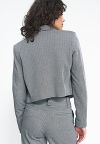Krótkim blazerze w szarym wzorze houndstooth, z wyraźnymi ramionami, klapami i gładką fakturą. Dopasowany krój z detalem na plecach.