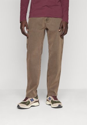 Mann trägt braune Hose mit geradem Schnitt, bordeauxfarbenes Langarmshirt und beige, grüne und blaue Sneakers, steht vor weißem Hintergrund.