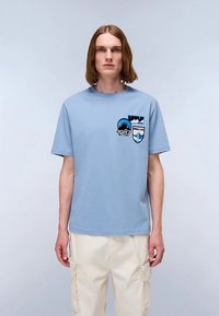 Light blue cotton t-shirt with short sleeves, featuring two patches: a mountain design and a shield, both in contrasting colors.