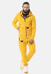 Gelbe Kapuzenjacke mit einer Vorderseite-Tasche und Reißverschluss, kombiniert mit passenden Hosen. Beide aus weichem Stoff, mit schwarzen Akzenten und Reißverschlüssen.