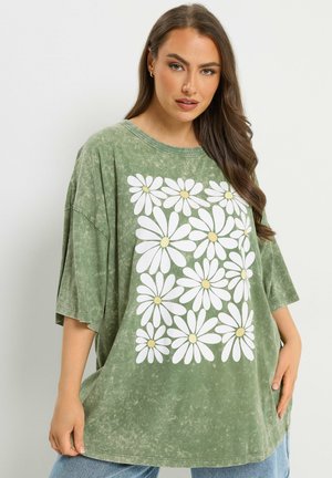 Frau mit langen braunen Haaren, die ein oversized ausgewaschenes grünes T-Shirt mit weißen Gänseblümchen und hellblaue Jeans trägt, steht vor einem einfarbigen Hintergrund.