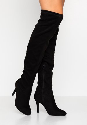 Bottes hautes noires en daim, à talons hauts avec fermeture éclair latérale, portées sur des jambes croisées sur un fond blanc uni.