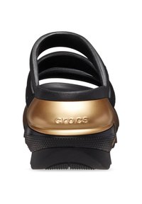 Zwarte klompschoen met een open achterkant, met een gouden hakaccent en een getextureerde rubberen zool. "Crocs" logo gedrukt op de hak.