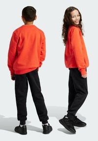 Sudadera naranja de corte relajado, combinada con pantalones jogger negros y zapatos negros. Cabello rizado y diseño simple, sin patrones.