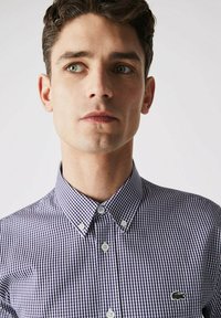 Camicia a quadri blu e bianca con colletto button-down. Presenta un piccolo logo verde sul lato sinistro del petto. Tessuto di cotone morbido.