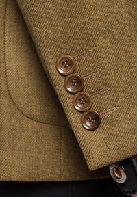 Rękaw brązowej kurtki w splot herringbone z wełny, z czterema okrągłymi, ciemnobrązowymi guzikami i widocznymi detalami szwów. Metalowy emblemat w pobliżu mankietu.