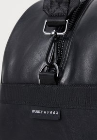 Sort rygsæk med en glat lædertekstur, toplynlås og en robust nylonrem med en metalclip. Mærket siger "MIN SPORT ER HYROX."
