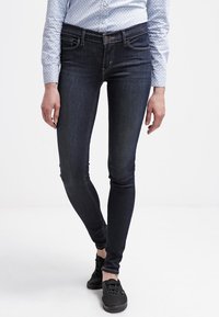 Persona con jeans ajustados oscuros, camisa de botones clara con estampado y zapatillas negras de pie contra un fondo blanco liso.