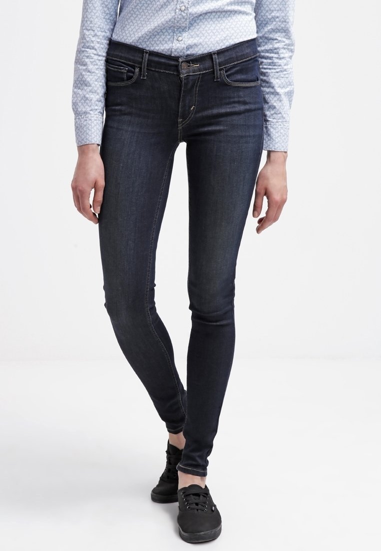 Persona con jeans ajustados oscuros, camisa de botones clara con estampado y zapatillas negras de pie contra un fondo blanco liso.