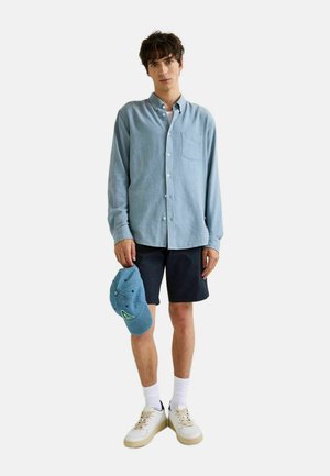Chemise à manches longues bleu clair à boutons, shorts noirs, chaussettes blanches et baskets crème. Tenant une casquette bleue avec un logo vert.