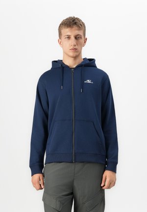 Ung mann iført en marineblå zip-up hettegenser med "O'Neill" logo og grå cargo bukser, stående mot en enkel hvit bakgrunn.