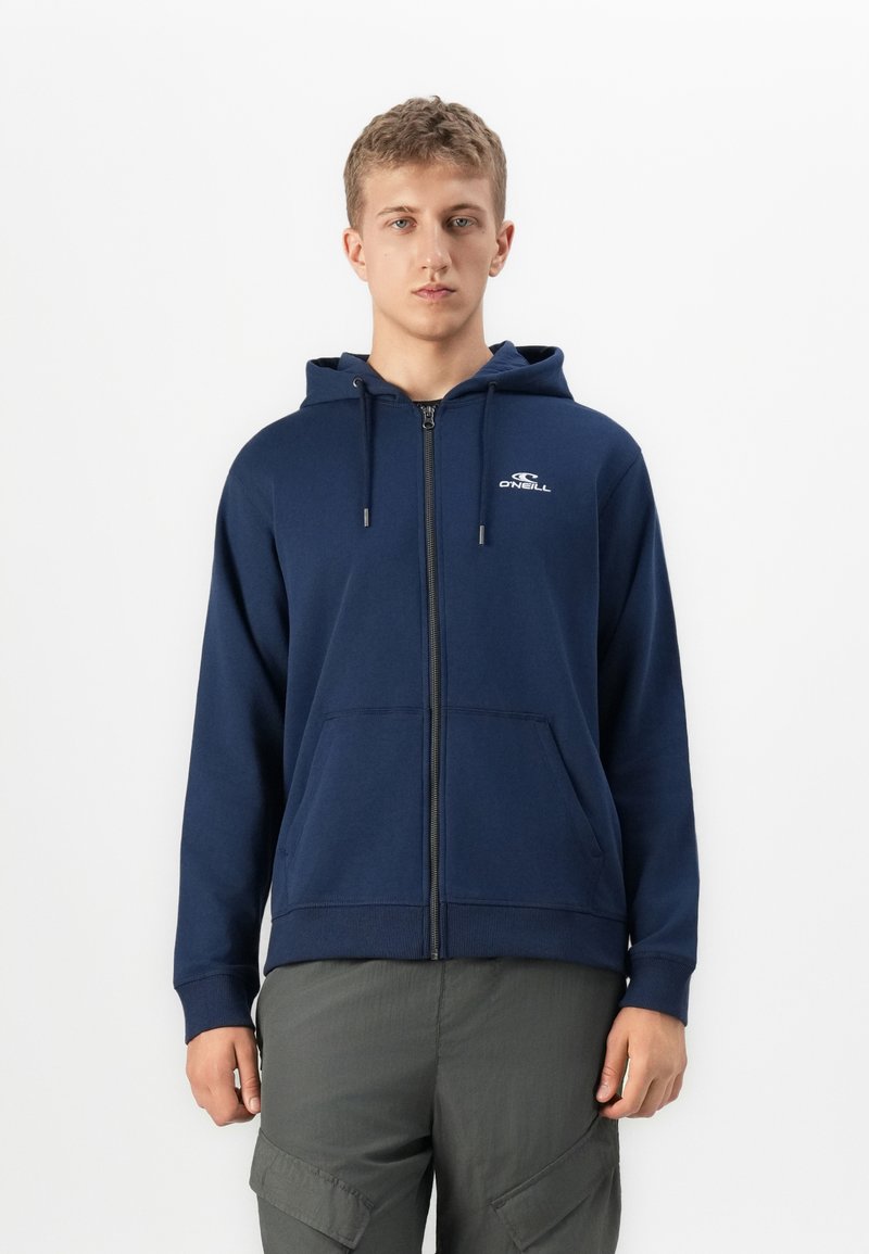 Giovane uomo che indossa una felpa con zip di colore blu navy con logo "O'Neill" e pantaloni cargo grigi, in piedi su uno sfondo bianco uniforme.