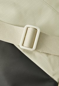 Tissu beige texturé avec une section noire lisse, présentant une boucle rectangulaire blanche maintenant une sangle. Matériaux durables, design pratique.