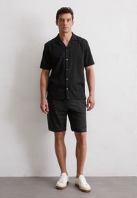 Zwarte kortemouwen overhemd met een gestructureerd design, gecombineerd met bijpassende donkere shorts en witte sneakers met tan kleurige zolen.