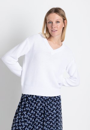 Weißer Strickpullover mit V-Ausschnitt, lockerer Passform, strukturiertem Stoff und geripptem Saum. Kombiniert mit einem gemusterten dunkelblauen Rock.