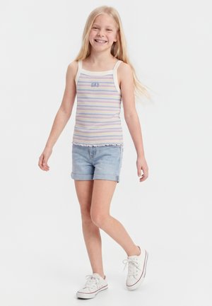 Χαμογελαστό κορίτσι με μακριά ξανθά μαλλιά, φοράει ριγέ αμάνικη μπλούζα, ανοιχτόχρωμα τζιν σορτς και λευκά αθλητικά παπούτσια, περπατάει με φόντο λευκό.