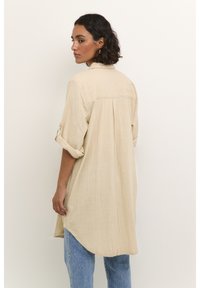Chemise en lin beige avec un dos long, des manches retroussées et un pli au centre. Associée à un jean bleu. Aucun motif visible ou quincaillerie.