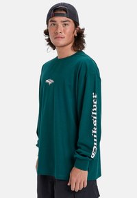 T-shirt à manches longues en teal, fabriqué en coton, avec un petit logo sur la poitrine et un grand texte blanc le long de la manche droite. Design à coupe décontractée.