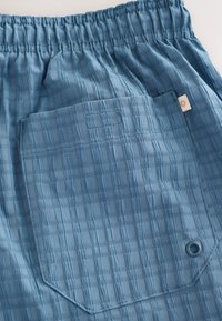 Siniset teksturoidut shortsit, jotka on valmistettu kevyestä materiaalista ja joissa on joustava vyötärönauha. Shortseissa on suuri päällystetasku ja koristeellinen merkki.