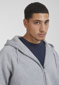 Gray zip-up hoodie with drawstrings, made of soft fleece material. Worn over a navy t-shirt, featuring a casual, relaxed fit and lightweight texture.