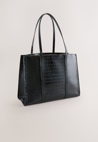 Czarna torba typu tote z tłoczeniem imitującym skórę krokodyla, z dwoma uchwytami, zamkiem błyskawicznym, prostokątnym kształtem, gładką fakturą oraz złotymi ozdobami.