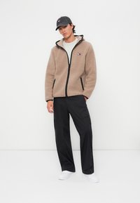 Giacca in fleece beige con zip, dettagli neri e logo ricamato; pantaloni cargo neri; sneakers bianche; cappellino grigio.