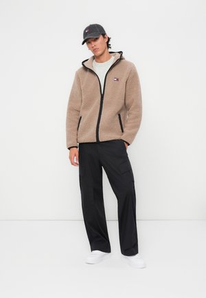 Chaqueta de forro polar beige con cremallera, detalles en negro y logo bordado; pantalones cargo negros; zapatillas blancas; gorra gris.