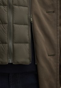 Olijfgroene gewatteerde jas met een gequiltde textuur, voorzien van een ritsluiting en een bruine buitenlaag. Functionele designelementen zichtbaar.