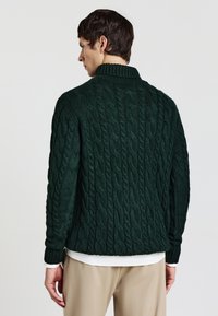 Dunkelgrüner Kabelstrickpullover mit hohem Kragen und gerippten Bündchen, der strukturierte Muster und einen lässigen Schnitt aufweist.