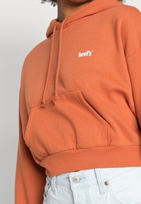 Orange cropped hoodie with a front pocket and drawstring hood; features a white Levi’s logo on the chest. Material appears soft and textured.