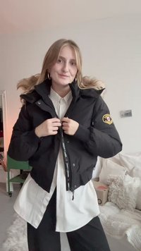 Mustan puffer-jakk on kapuutsiga, millel on varrukal logo lapiga ja esitasku tõmblukk. All on nähtav valge kaelusega särk.